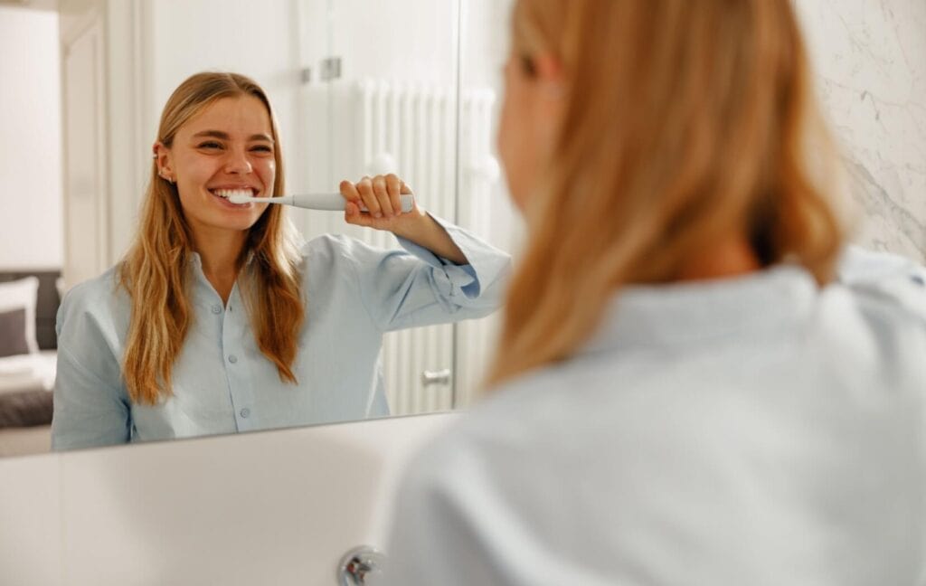 A person smiles in their bathroom mirror as they use an electric toothbrush to gently clean their teeth.
