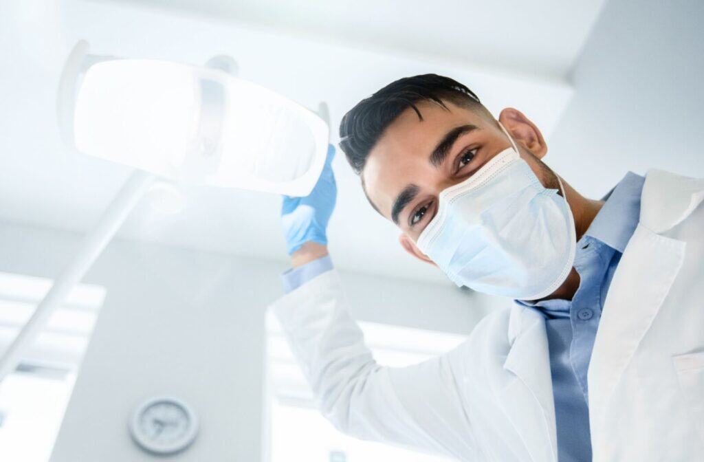 A patient's view of a dentist wearing a surgical mask and gloves adjusting a light in a clinic above a patient chair.
