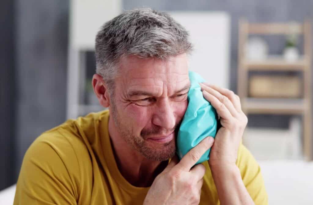 A person with grey hair sitting down and holding a turquoise ice pack to their left cheek.
