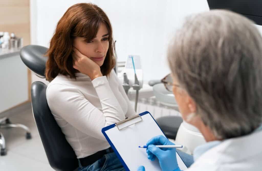 A woman holding her hand to her cheek while sitting in a dental chair. A dentist with grey hair takes notes on a clipboard.
