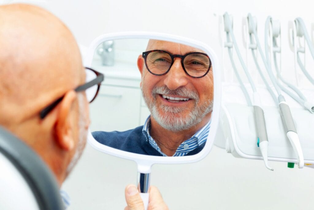 Older man wearing glasses smiling while looking at his reflection in a handheld mirror during a dental appointment.