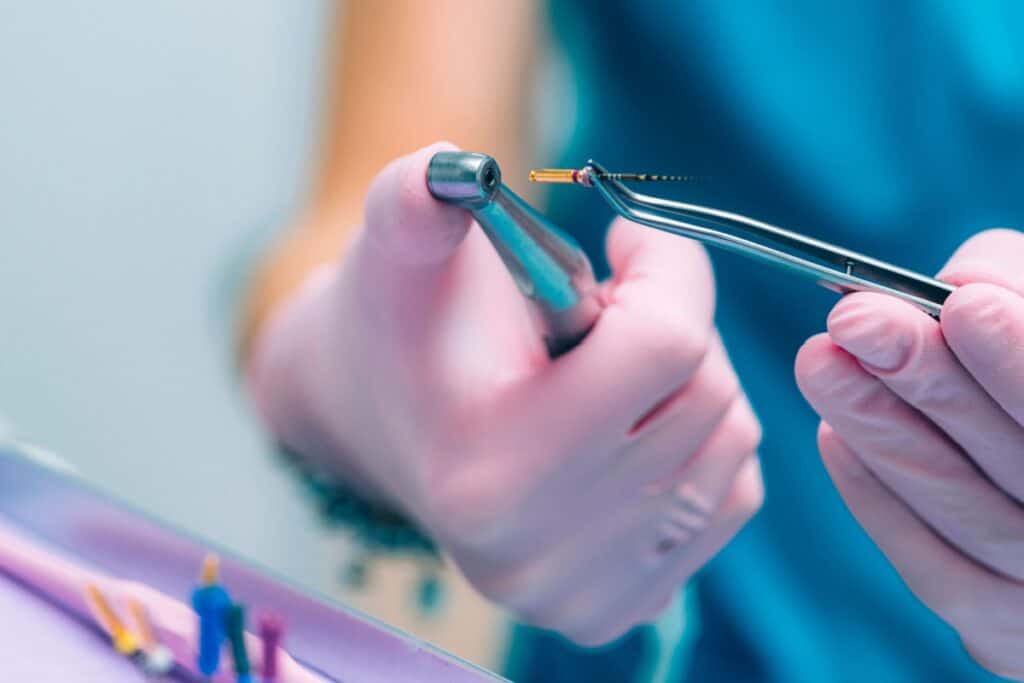 Gloved hands holding a dental drill and a small endodontic file, preparing instruments used in root canal treatment.