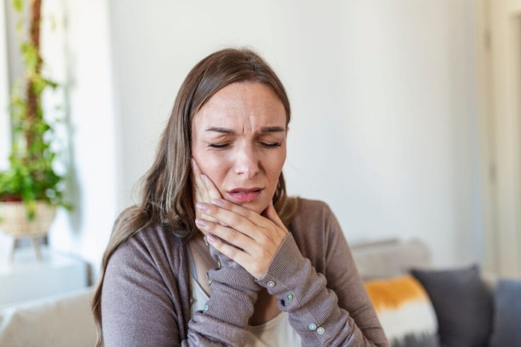 A woman indoors holding her jaw with both hands and wincing, indicating discomfort or possible toothache.