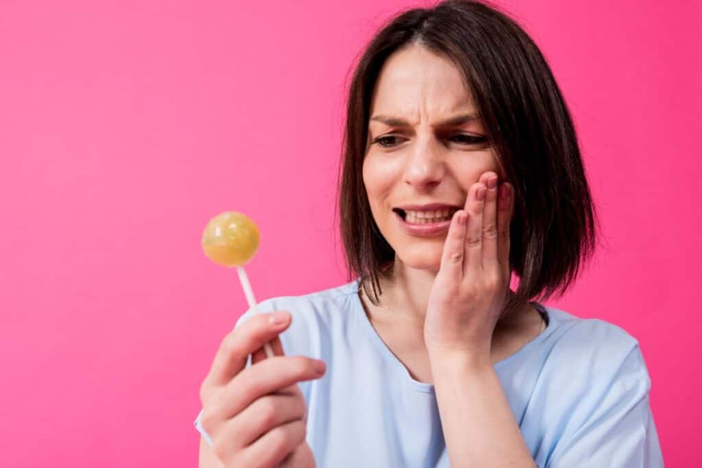 A woman holding a lollipop while touching her cheek with a pained expression, suggesting tooth sensitivity or dental pain. \