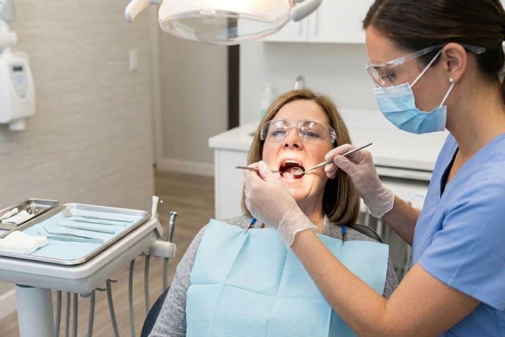 A dental hygienist wearing gloves, a mask, and protective glasses examines a patient’s mouth in a dental office while the patient reclines in a chair with a bib on.