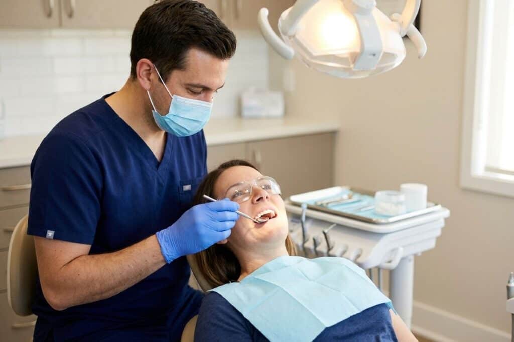 Dentist examining teeth of a patient reclining in a chair, using a mirror tool under an overhead light.
